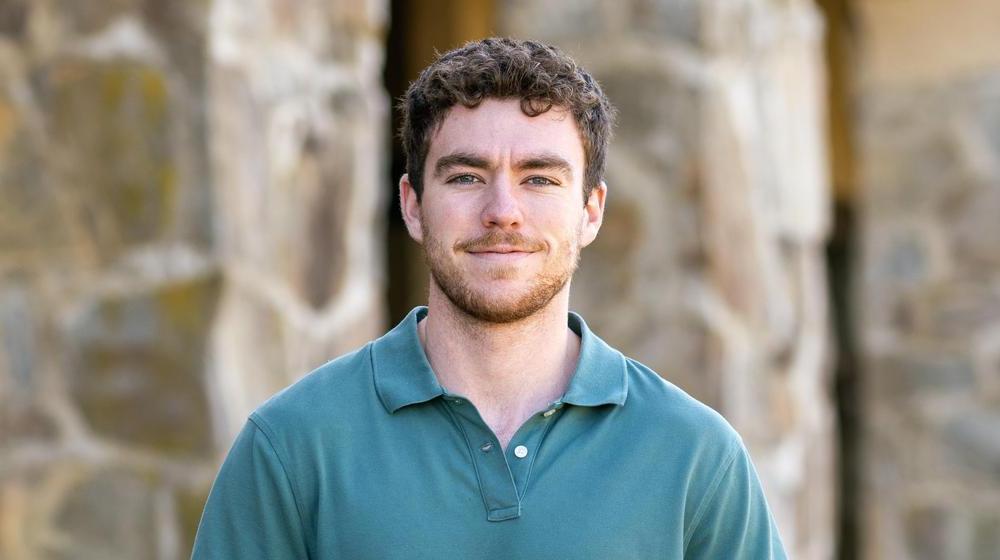 Young man with curly hair and a slight smile, wearing a green polo shirt, standing in front of a stone building.